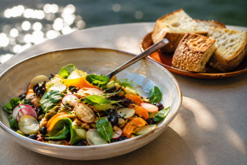 AI illustration: Close-up of a colorful light salad and small shared bread plate on a softly lit waterside dining table