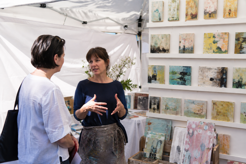 An adult artist speaking with a shopper beside a display of original small abstract paintings under a white outdoor market canopy