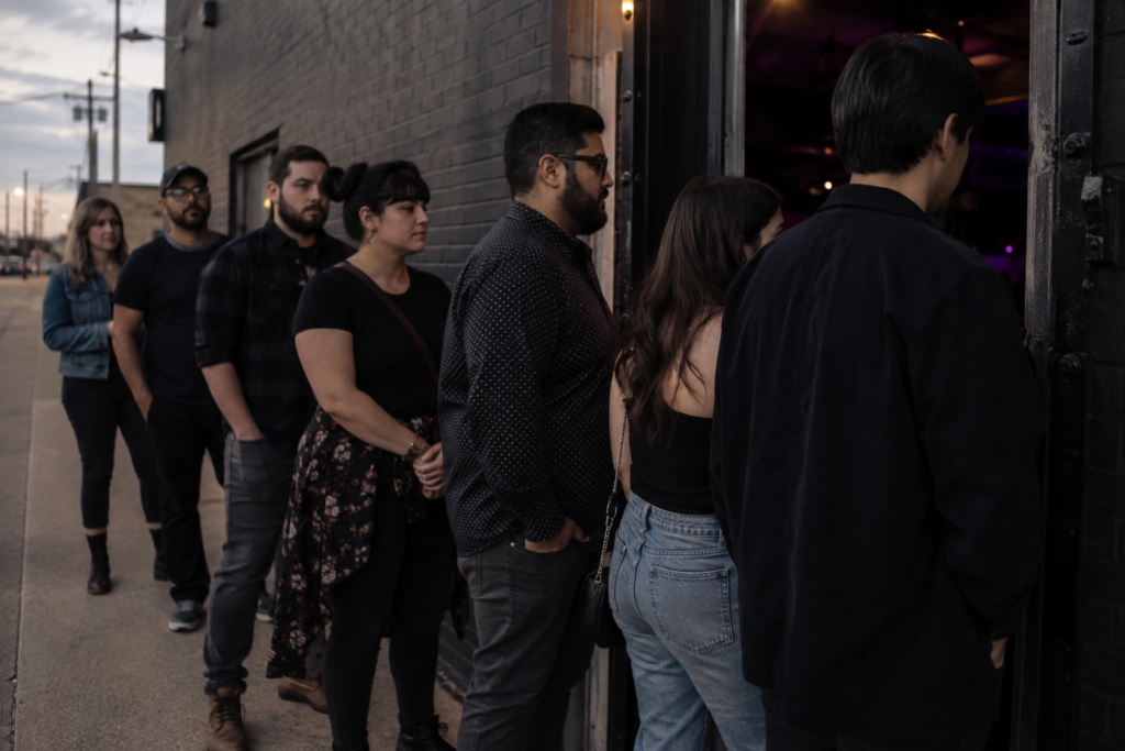 A compact line of adults waiting politely at a generic music venue doorway at dusk