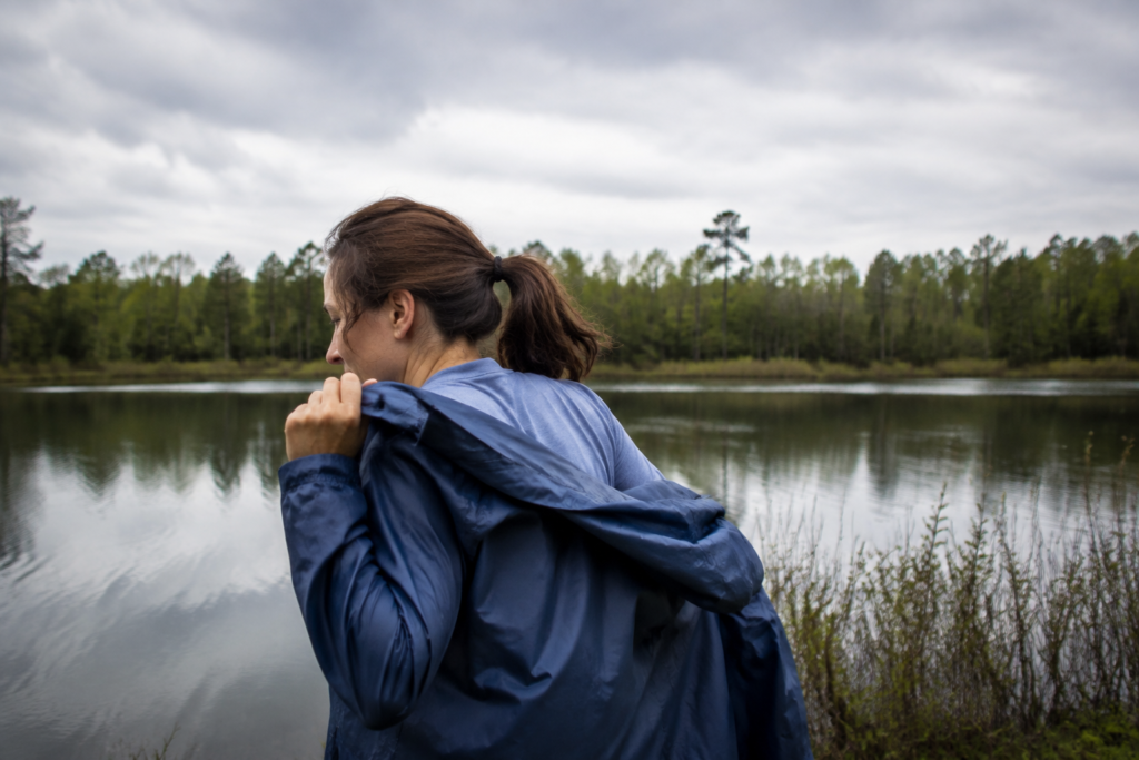 A visitor pulling on a lightweight rain jacket beside a calm small lake under cloudy skies