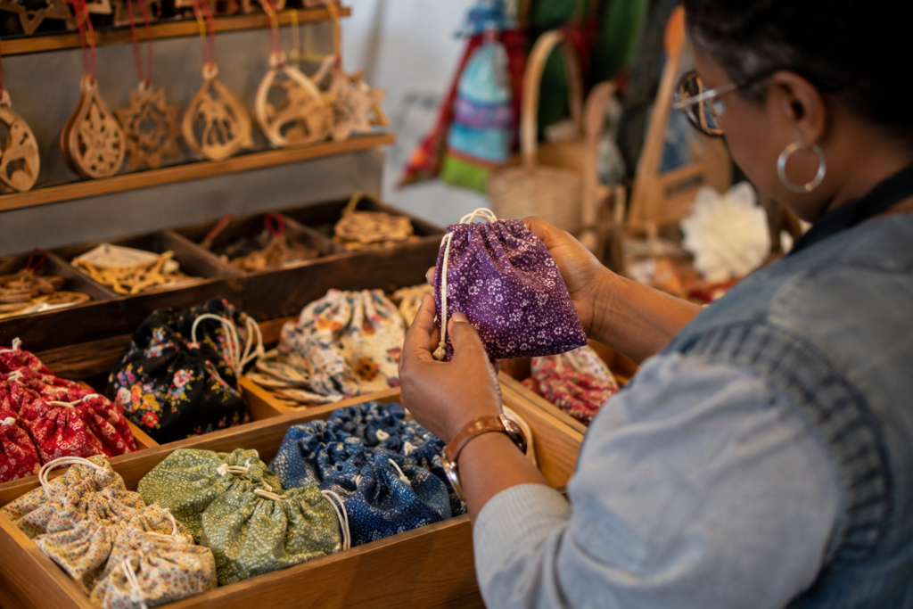 Close detail of a shopper examining hand-sewn fabric gift pouches and locally crafted wooden ornaments inside a bright indoor market