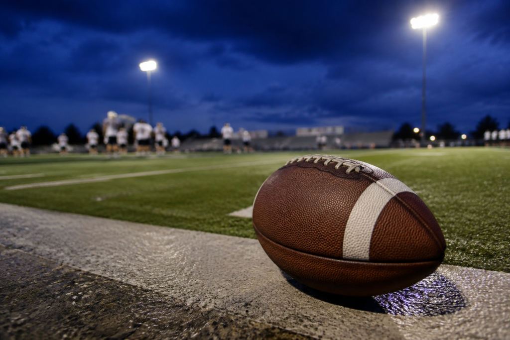 Close dramatic brown American football resting on the sideline of a community football field under evening stadium lights