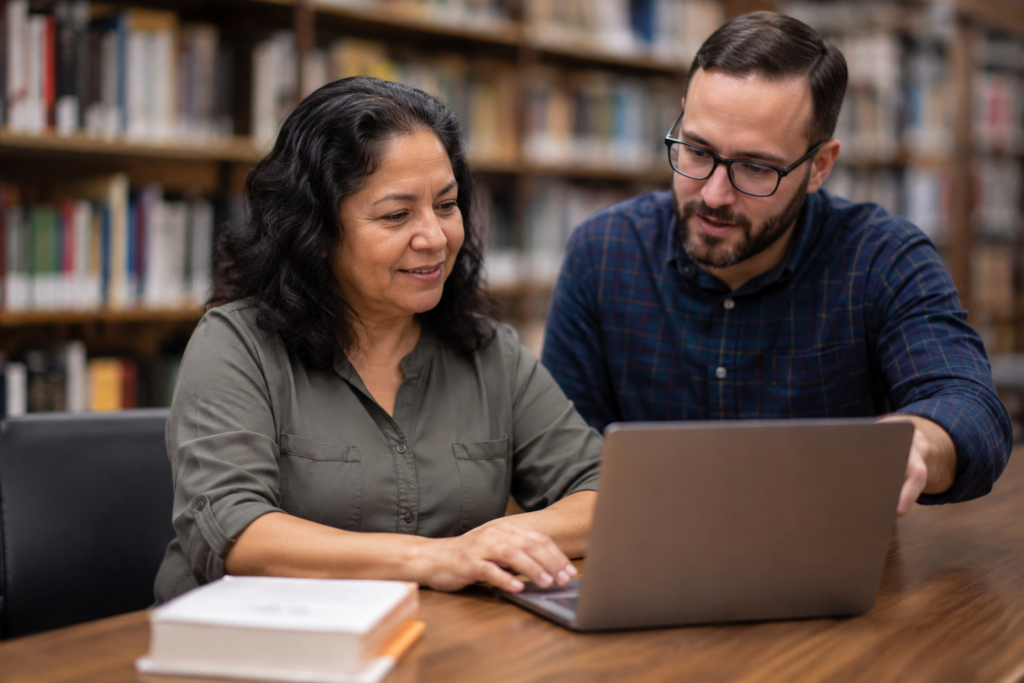 Adult learner practicing on a library laptop with instructor beside them