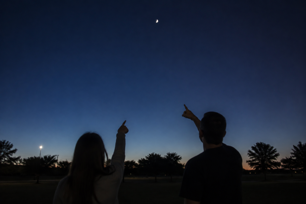 Two adults pointing up toward a modest half moon in a clear night sky at a dark park